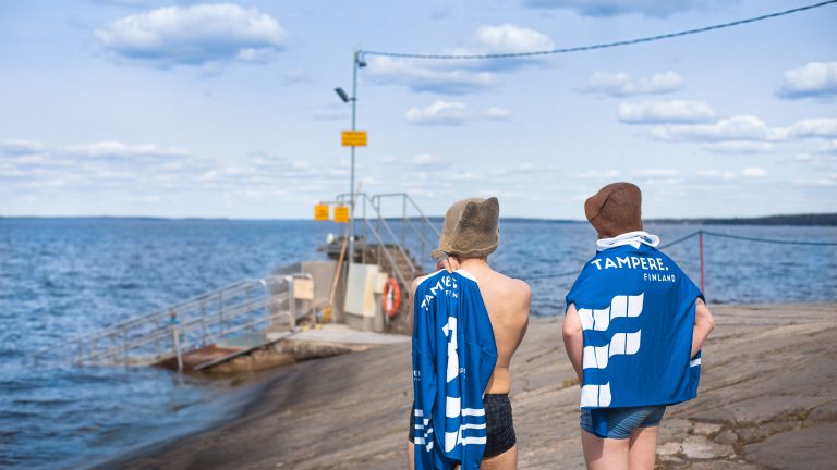 Miehet seisovat uimahousut päällään Rauhaniemen kylpylän kallioilla siniset Tampere.Finland-paidat harteillaan ja katselevat Näsijärvelle.